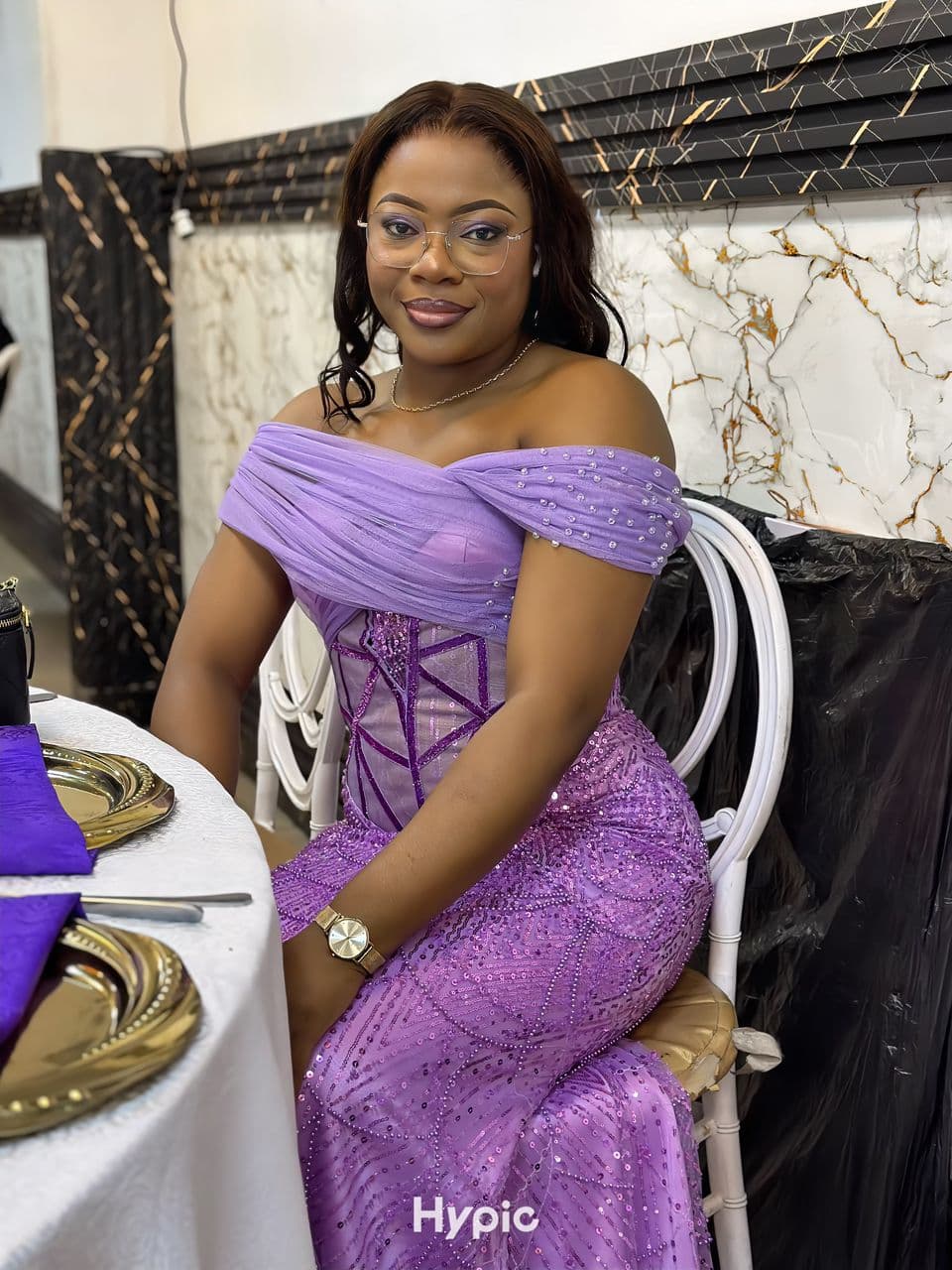 Femme souriante en robe violette à paillettes assise à une table de réception élégante.