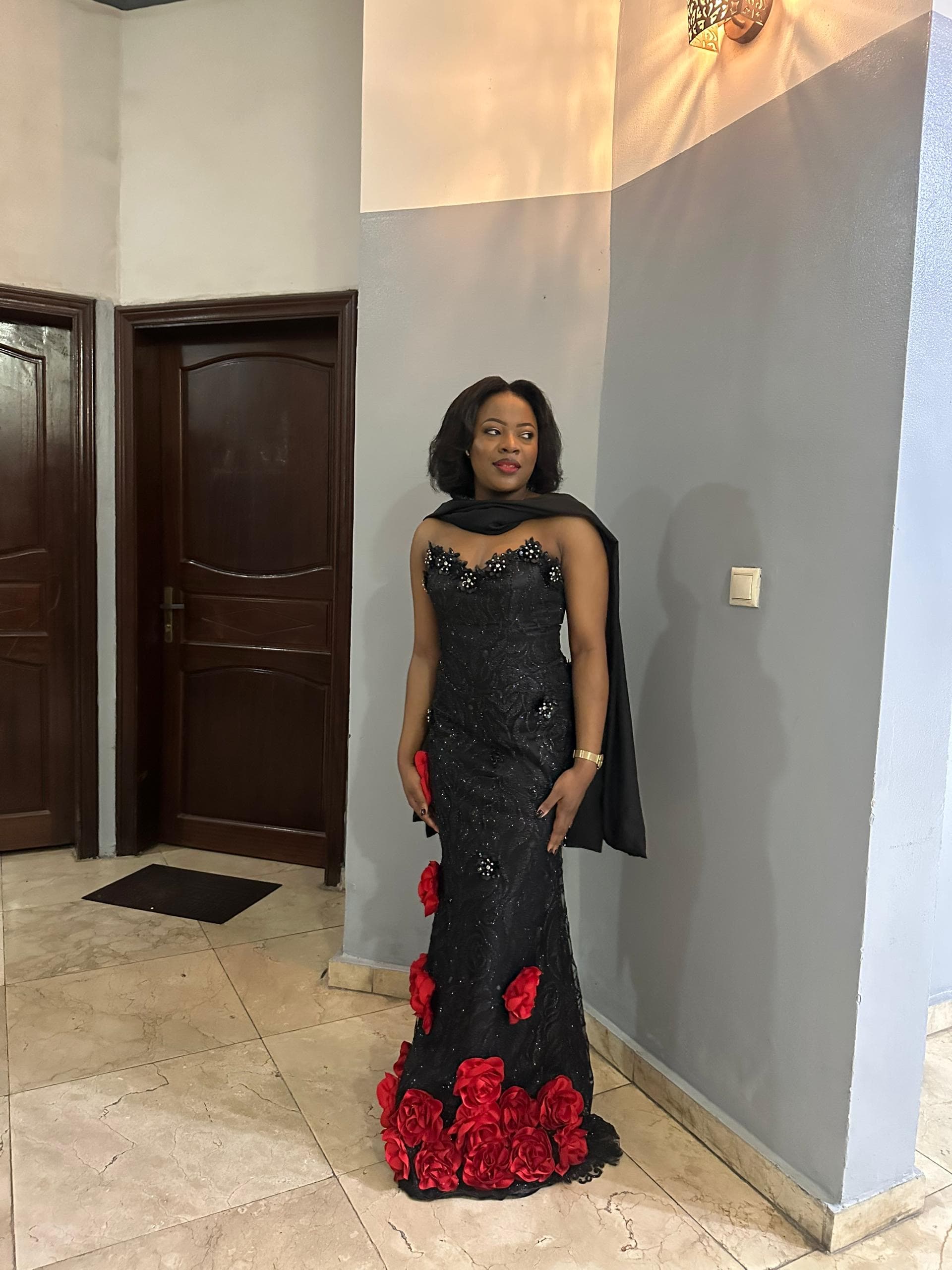 Femme élégante en robe de soirée noire ornée de roses rouges posant dans un couloir.
