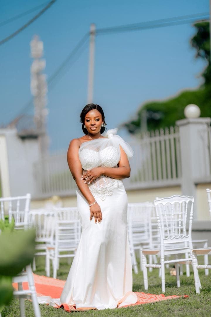 Femme en robe blanche élégante posant dehors parmi des chaises blanches sur un tapis rouge.