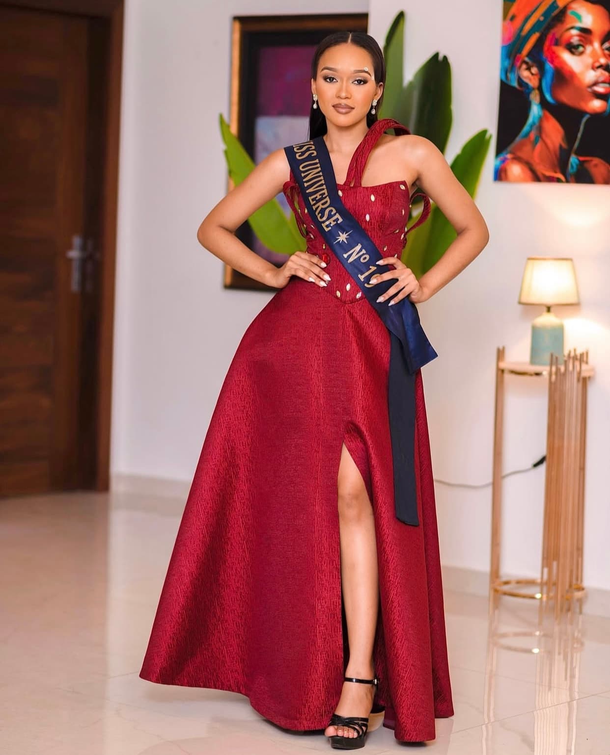 Miss Universe en robe rouge fendue, posant avec élégance les mains sur les hanches.