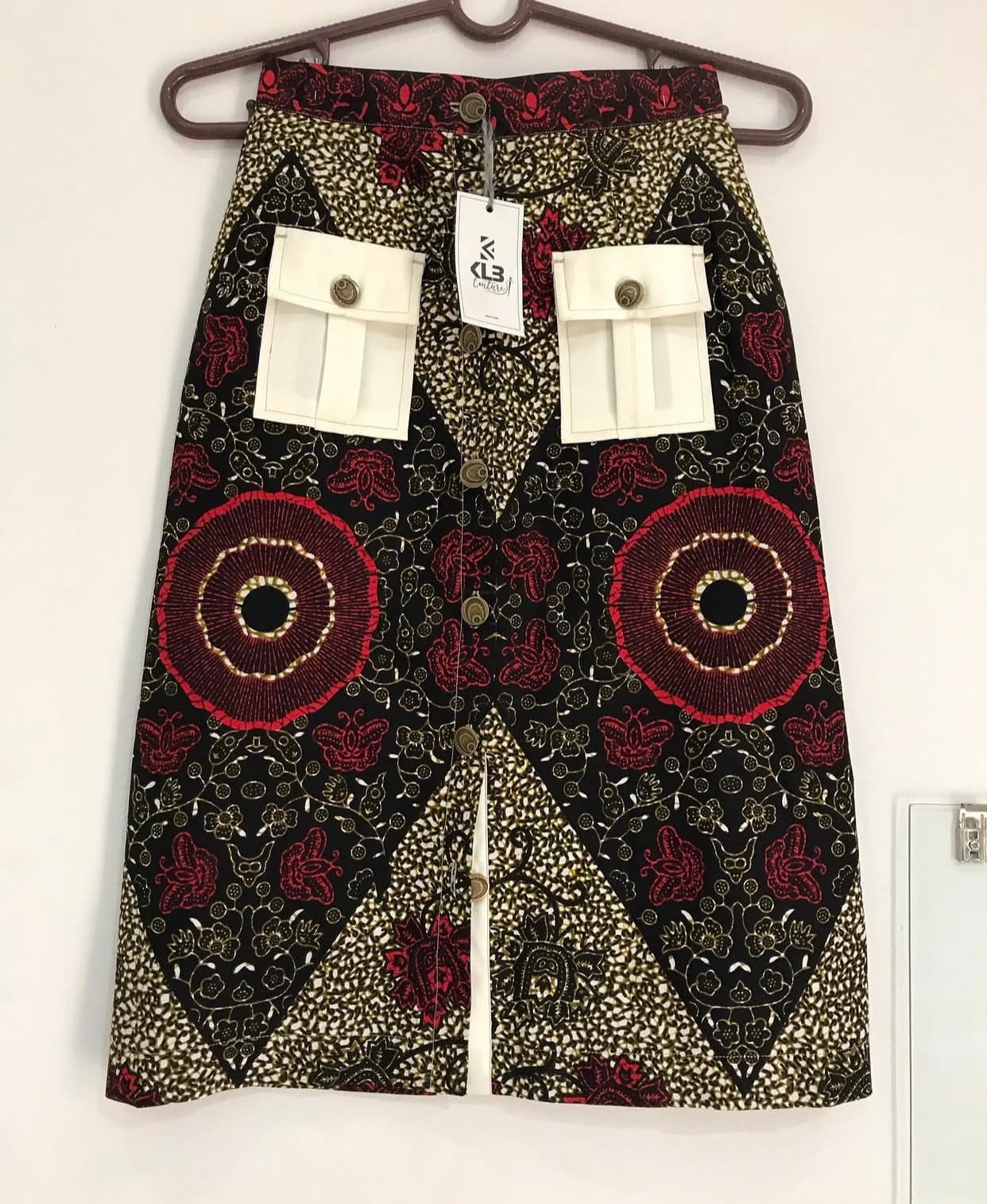 Jupe à imprimé africain avec motifs rouges, poches blanches et boutons, suspendue sur un cintre.