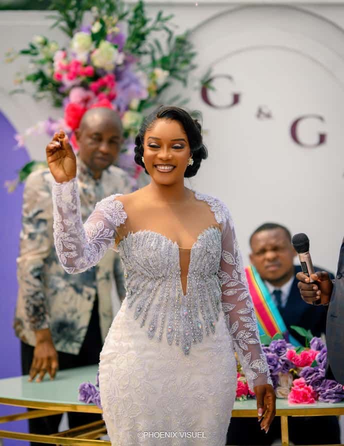 Femme souriante en robe de mariée blanche perlée levant la main lors d'une cérémonie.